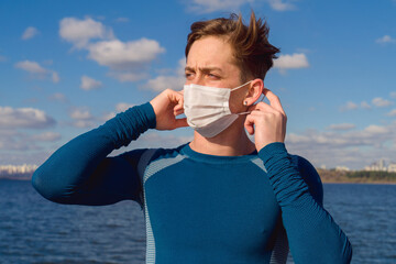 Male model posing with mask by the sea on a sunny autumn day. Image of handsome caucasian young man wearing a mask under blue sky with sportswear. Avaible copy space area. 