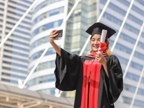graduated woman in graduation gowns holding diploma, making selfie photo with mobile phone. City building background. Education  and successful concept.