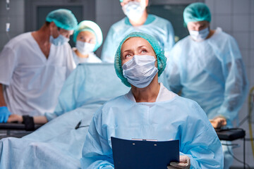 portrait of assistant of doctors making operation to patient, woman in mask and uniform hold digital tablet of patient diagnosis, check, looks up. while colleagues surgeons do operation in background