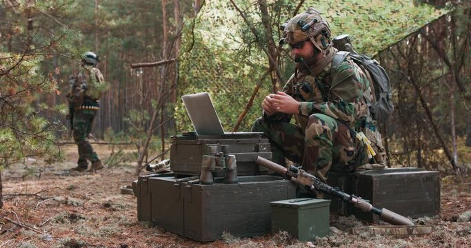 A bearded commander in a military uniform, in a tactical vest with a helmet on his head, chatting online on a laptop at a temporary forest base. In the background you can see a soldier guarding the