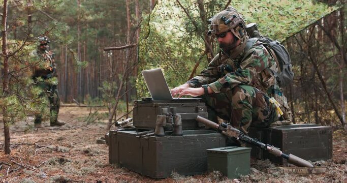A bearded commander in a military uniform, in a tactical vest with a helmet on his head, types on the keyboard of a laptop in a temporary forest base.