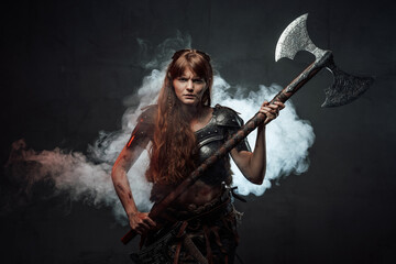 Violent and savage scandinavian female warrior holding huge two handed axe in dark foggy background.