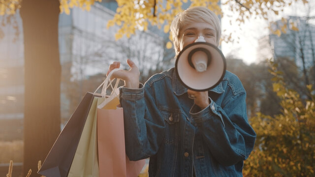 Young blond woman with shopping bags announcing sales season with loudpeaker in the city park in autumn. High quality photo