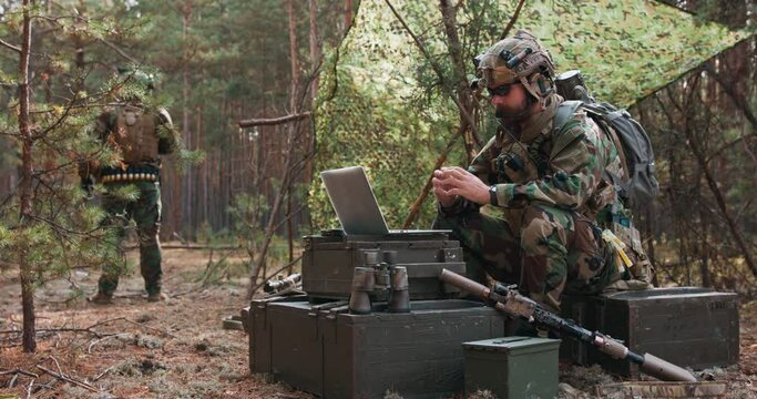 A bearded commander in a military uniform, in a tactical vest with a helmet on his head, chatting online on a laptop at a temporary forest base. In the background you can see a soldier guarding the