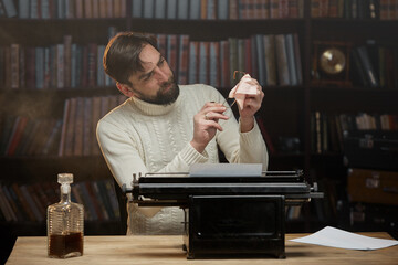 A focused male writer with glasses is typing a novel on a typewriter. A man with a beard in a white sweater wipes glasses sitting at the home table, in the background of the library