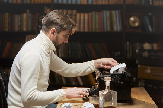 A focused male writer with glasses is typing a novel on a typewriter. A man with a beard in a white sweater using a retro typewriter sitting at a home table in the background of the library