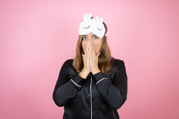 Young caucasian woman wearing sleep mask and pajamas over isolated pink background with her hands over her mouth and surprised