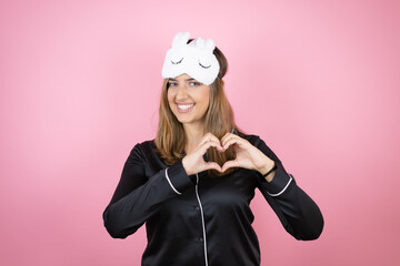 Young caucasian woman wearing sleep mask and pajamas over isolated pink background smiling in love showing heart symbol and shape with hands