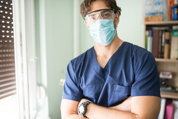 Young brunette medical doctor wearing a protective face mask and protective glasses and blue lab coat standing in the hospital