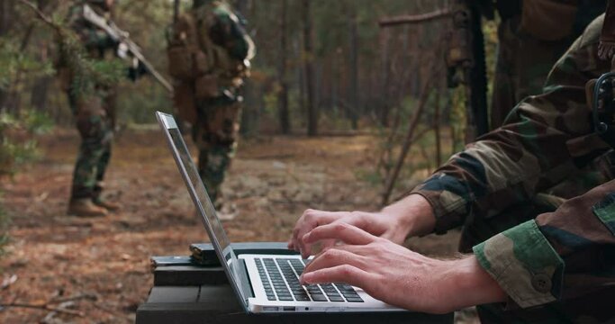 Portrait of bearded commander in a military uniform, in a tactical vest with a helmet on his head, typing on the keyboard of a laptop in a temporary forest base. In the background you can see a