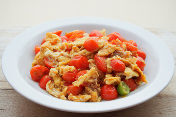 Tomato scrambled eggs in white ceramic dish on wooden table against blurred background with copy space.