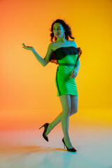 Fototapeta na wymiar Dance. Beautiful girl in black green fashionable, romantic outfit on bright gradient yellow-orange background in neon light. Full-length portrait. Copyspace for ad. Summer, fashion, emotions concept.