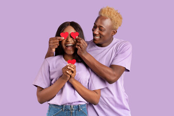 Funky black guy covering his girlfriend's eyes with red hearts, surprising her on lilac studio background