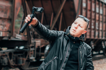 Fototapeta na wymiar Handsome Guy in Black Clothes and Sunglasses Making Selfie or Streaming Video Using Action Camera with Gimbal Camera Stabilizer at Railway