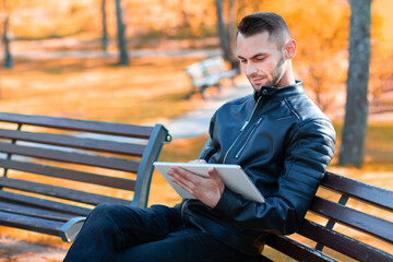 Fototapeta premium Handsome Young Man Sitting on the Bench and Using Big White Tablet PC at the Beautiful Autumn Park