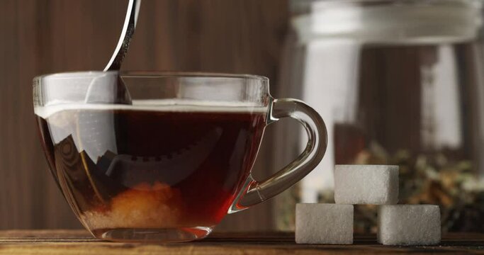 Stir sugar with a spoon in a clear glass Cup with hot tea closeup, slow motion. Next to it are pieces of refined sugar. A can of hand-picked herbal tea stands against a wooden background. Tea ceremony