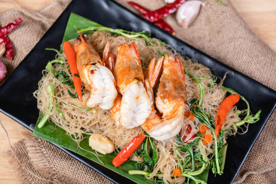 Shrimp, noodlesrice noodle and butter garlic fried shrimps sprinkled with pieces of chilli and chopped parsley on white dish on natural old wooden planks with spaghetti tongs, view from above, food
