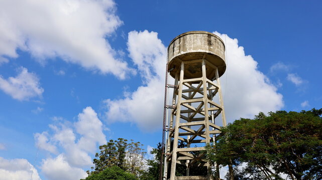 Old concrete water tank on the tower. A large water reserve tank for the community below the green trees. On the sky background there are white clouds with a copy space. Selective focus - Powered by Adobe
