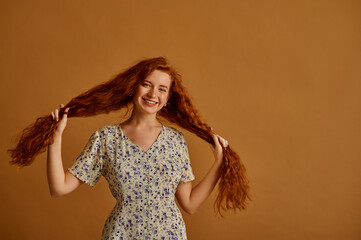 Happy smiling redhead woman with long natural curly hair wearing floral print summer dress, posing on beige background. Model holding her hair. Haircare conception. Copy, empty space for text