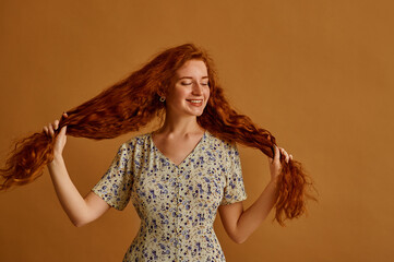 Haircare conception: happy smiling redhead woman with long natural curly hair holding it.  Model wearing floral print summer dress, posing on beige background. Copy, empty space for text