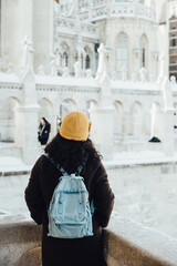 Back view of unrecognisable young woman tourist in winter coat and yellow hat with blue backpack looking at gothic architecture of Fisherman Bastion in Budapest, Hungary during bright winter day in De