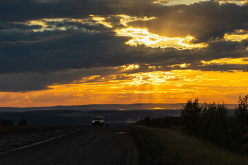 Black silhouettes of trees and a gray road are on a blue sky background with orange and yellow sunset