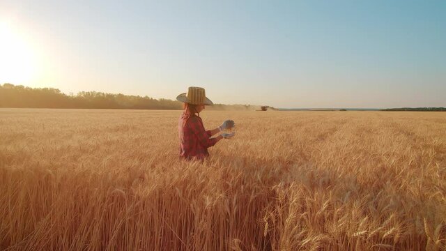 Young woman farmer in hat standing against the background of a working combine harvester at sunset. The concept of agricultural work and harvesting.