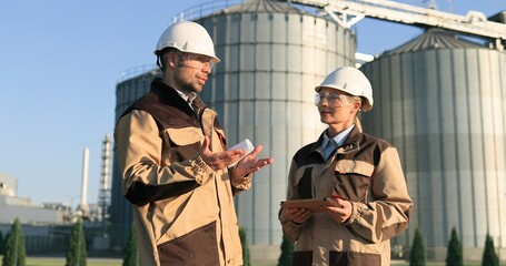 Portrait of Caucasian male and female builders in helmets with tablet and graphs speaking outdoors. Woman and man engineers in goggles discussing industry manufacturing plan. Engineering concept