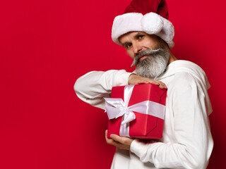 Senior bearded man holding a gift box on a red background and looking at the camera with a smile. Stylish and fashionable Santa Claus wishes you a Merry Christmas and a Happy New Year 2021.