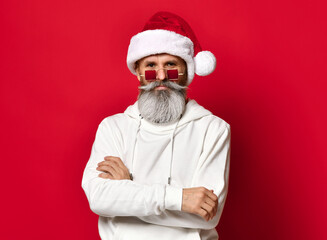 Fototapeta na wymiar Winter fashion and sales. Portrait of a handsome senior man in a Santa hat, winking while standing against a red background. The hipster is holding glasses and looking at the camera. Place for text.