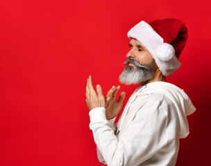 Fototapeta na wymiar A bearded man in a Santa hat stands with his eyes closed and prays hopefully, desiring something. The concept of human emotions and desires for new year. Red background. snow time.