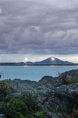 mountains and lights with clouds in the background and rock in the front and ocean in the middle