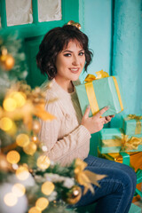 A girl in a white sweater sits on the porch near the door against the background of a Christmas tree with a gift. New Year's holidays. Surprise box