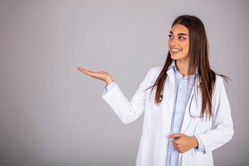 Smiling woman doctor showing aside to copyspace blank place. Confident female medical worker portrait pointing with finger to left side at empty space in studio isolated on gray background