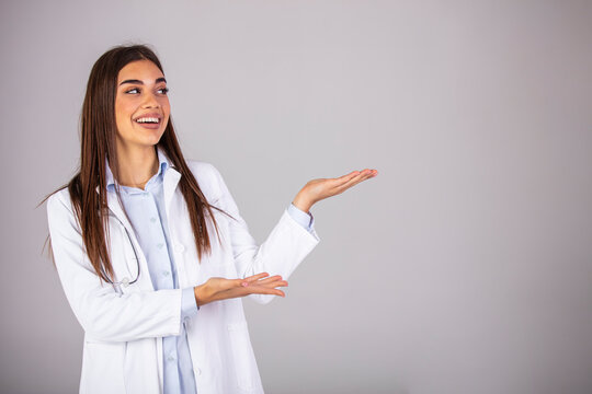 A happy doctor gesturing towards the side - copyspace. Beautiful smiling young woman in white coat pointing at copy space and looking. Waist up studio shot on gray background.