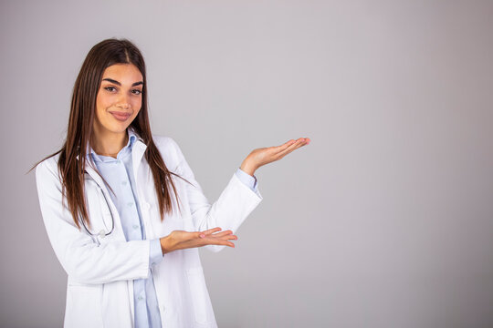A happy doctor gesturing towards the side - copyspace. Beautiful smiling young woman in white coat pointing at copy space and looking. Waist up studio shot on gray background.