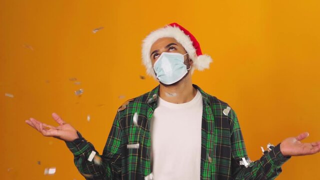 Sad man in Santa hat and medical mask standing under falling confetti against yellow background