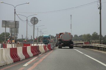 Milk truck with a gray barrel drives along a narrowing road, road works on a suburban highway on a summer day, rear view on gray sky background