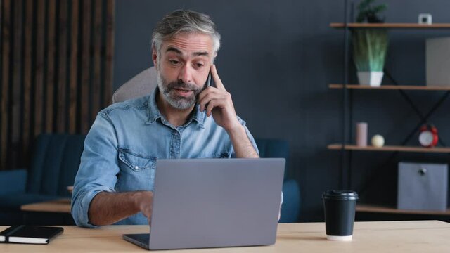 Matured Freelancer, Businessman is very Busy, is talking on his Phone, working on Laptop at the same Time. Handsome Bearded Male is working indoor at Home Office. Easy Technology. Freelancer.