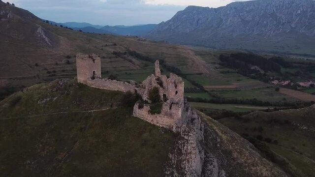 Aerial view approaching the ruins of a medieval fortress (Cetatea Trascaului) that sits on top of a steep hill with mountains in the background