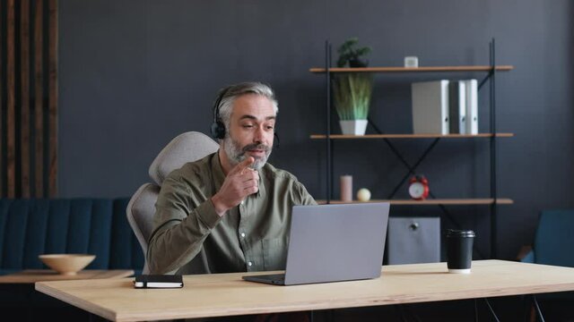 Confident Successful Mature Businessman, Mentor, Coach, Teacher holding Video-Call Class. Bearded Man is wearing a Headset, holding a Seminar, Conference, Webinar. Online Job, Indoor. Using Laptop.