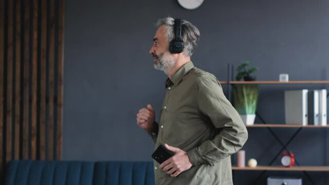 Portrait of handsome Man in Headphones is listening to Music and Dancing Funny. Mature Bearded Man is having fun while dancing in his Flat. Having fun alone Indoor. Spending Time at Home. Funny Man.