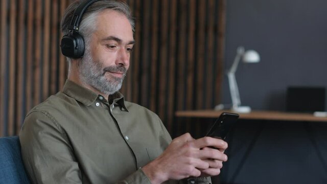 Satisfied Bearded Man is relaxing on the Couch at Home, chatting with Friends on Smartphone, browsing Social Networks. Mature Man wearing big Headphones. Casual Surfing Lifestyle. Spending Time Home.