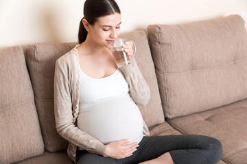 Pregnant woman with a glass of water sitting on sofa at the home