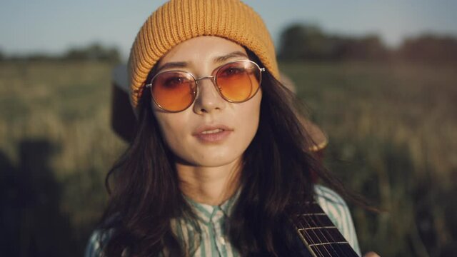 Beautiful Asian hipster woman gutar player walking on a sunny field with a acoustic. The girl in trendy sunglasses enjoy summer sunset and walking forward in countryside. Youth and creativity concept
