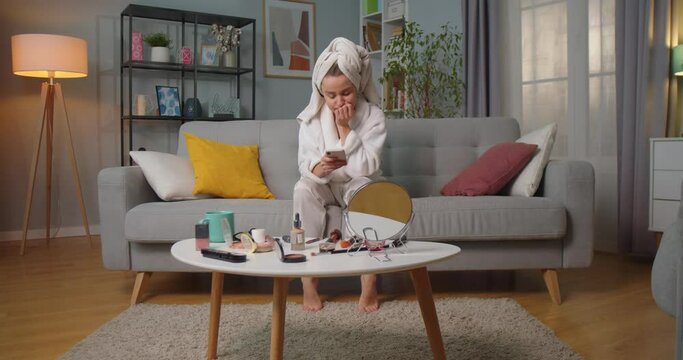 Middle plan of woman in bathrobe and towel sitting on sofa in living room texting, chatting, reading messages using smartphone. Makeup kits on table in living room. Concept of lifestyle, self-care