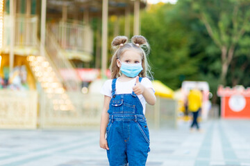 a baby girl stands in an amusement Park wearing a protective medical mask and gives a thumbs up