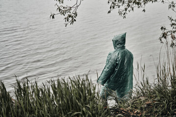 A man in a green raincoat on the shore near the water. Mainly cloudy