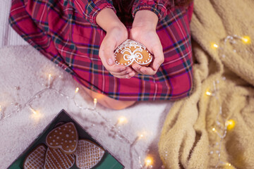 Homemade heart-shaped gingerbread cookie in the hands of a little girl. Red plaid dress, sitting on the floor.