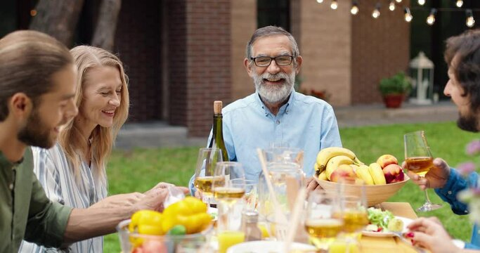 Caucasian adults kids visiting senior father or grandfather on weekend and having dinner on fresh air at back yard. Family eating and talking outside. Celebrating.